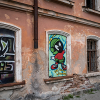 The image reveals a weathered building reminiscent of an old Latvian fortress, adorned with three large windows covered in colorful graffiti. The left features a stylized symbol, the center displays a cartoon character in a red and green outfit, and the right has a portrait of a bald man with glasses.