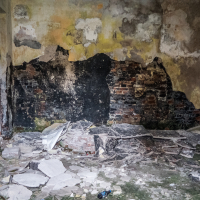 A dilapidated room within Daugavpils Fortress, featuring peeling paint and a crumbling ceiling. The walls are marked with black mold and exposed brick. Debris covers the floor, with broken plaster and dirt scattered about. A partially open door on the left hints at further decay and neglect beyond.