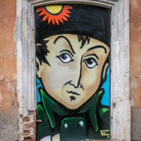 Street art painted on a window depicts a cartoonish man in a dark coat and hat with a sunflower above his head, reminiscent of Daugavpils' vibrant spirit. The wall surrounding the window is weathered and peeling, adding texture to this Latvian-inspired scene within its rustic white frame.