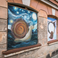 Street art on a weathered building in Daugavpils fortress shows two paintings. The first depicts a snail with a spiral shell against a starry night, echoing Van Gogh's style. The second features a stylized snail with a geometric spiral shell. Surrounding walls have textured, peeling paint.