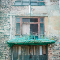 An old, weathered brick building facade with arched windows in Daugavpils, Latvia. The structure resembles a fortress, partially covered in green netting. The lower section has large, worn doors and a small canopy with visible vegetation growing on it. The wall and windows show signs of decay and aging.