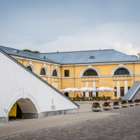 A yellow and white neoclassical building with large arched windows sits under a cloudy sky at the edge of Daugavpils Fortress. A row of umbrellas shelters outdoor seating in front, surrounded by potted plants with colorful flowers. Wide steps lead up to the entrance on the right, and a paved walkway runs alongside.