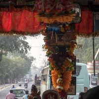 A bus interior decorated with colorful garlands, statues, and ornaments hanging above the dashboard. Through the front window, a busy street in India is visible with vehicles and motorbikes. A red curtain and various posters adorn the upper part of the windshield. Trees line the road outside.