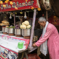 An elderly man in a pink traditional attire stands beside a street food stall named "Agga Anda Bhandar." The stall showcases various vegetables, including cauliflower and eggplants, along with metal containers and loaves of bread. Yellow decorative elements hang from the roof, evoking a vibrant scene typical of India.