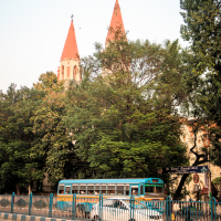 A scenic cityscape reminiscent of a vibrant street in India features a blue bus driving past. In the background, tall green trees partially obscure a building with two distinct red conical towers. The street is bordered by a blue and white striped fence, and a clear sky overhead hosts a few flying birds.