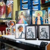 A display featuring various religious merchandise, including framed photographs and sketches of Mother Teresa, figurines of religious figures, and framed art of Jesus and the Virgin Mary. Also included are souvenir items like mugs, many reflecting India's rich spiritual heritage. All items are arranged on a well-lit white surface near a glass counter.