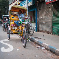 A man is sitting on a yellow cycle rickshaw parked on the side of a narrow street in India, with closed shops and green rolling shutters. Other rickshaws and pedestrians are visible in the background. The street is marked with white paint, giving the urban area a slightly cluttered appearance.