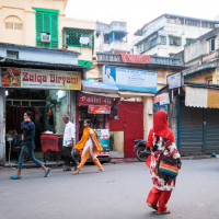 A street scene in an urban area of India, with several people walking past small shops. One shop sign reads "Zaiqa Biryani," another reads "Sindhu Cardio Clinic." A woman in a red headscarf and colorful outfit walks by, while two individuals on a blue scooter ride past. The buildings show signs of weathering.