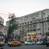 A bustling city street scene with multiple yellow taxis, cars, and pedestrians evokes the vibrant chaos of India. The backdrop features a large, grey, historic building with numerous windows and an array of colorful shop signs at ground level. The sky is clear with daylight fading; trees and power lines are visible.