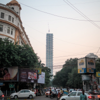 A bustling urban street intersection in India teems with people and vehicles, including cars and motorcycles, under a clear sky. A tall, modern skyscraper under construction is visible in the background, while a historic building with an ornate façade stands on the left. The street is lined with trees and various signs, including blue barriers and billboards.