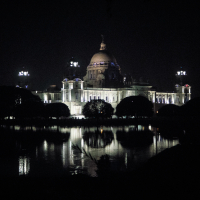 The majestic Victoria Memorial in India, illuminated at night, stands tall with its grand dome and intricate architecture. Reflections of the memorial shimmer in the tranquil water in the foreground, while trees frame the lower part of the structure against the dark sky.