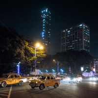 A nighttime city scene with multiple yellow taxis and cars at an intersection. Streetlights illuminate the area, and tall buildings with bright lights, including one with a distinctive blue vertical light pattern, are visible in the background. Trees line the streets, adding some greenery reminiscent of bustling cities in India.