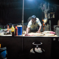 A person under a dim light works at an outdoor food stall in India. The stall features a table with several ingredients, including a bowl of noodles, condiments, utensils, and a gas cylinder beneath. A dark backdrop with hanging bags and tools forms the background, creating a rustic ambiance.