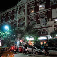 Night scene in front of a historic, red and white multi-story building adorned with arches, balconies, and intricate designs reminiscent of India. Street is lined with parked cars and lit by streetlights. People walk on the sidewalk near brightly lit storefronts, one labeled 'Fire and Ice.' Trees partially obscure the façade.