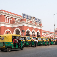 The image shows a lineup of green and yellow auto rickshaws parked in front of a historic red brick building with arched windows and white accents. The building, marked "Agra Fort," is likely a railway station in India. Several people are near the rickshaws or walking by. A streetlamp is also visible.