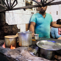 A man in a teal shirt stands by a rustic kitchen stove with multiple pots and pans, some emitting steam. The stove, constructed from aged, rough materials, has visible flames. Another man sits in the background against tiled walls. The setting appears to be an old-fashioned eatery in India.
