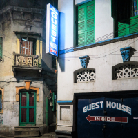 A dimly lit street corner at night features old buildings with green shutters and balconies, reminiscent of colonial-era India. A neon sign on the left reads "Guest House," while a larger blue and white sign on the right says "Guest House in side" above a garage door. Electrical wires crisscross overhead.