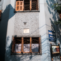 The light blue building with brown shutters, adorned with a statue above three windows, stands in India. A sign reads "Missionaries of Charity Mother House," while another on the right offers more details. Shadows from nearby trees dance across the wall, creating a tranquil scene.