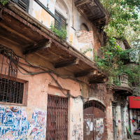 A weathered, multi-story building reminiscent of old India with peeling paint and overgrown plants on the upper sections. The facade features arched windows with shuttered blinds. A gated doorway is flanked by posters and graffiti, while nearby foliage partially obscures a corner balcony above the tiled sidewalk.