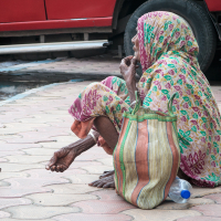 An elderly person, sitting on a tiled sidewalk in India, wears a colorful, patterned outfit and headscarf. They are barefoot, with a cloth bag beside them. A stainless steel container and plastic bottle rest nearby, while a red vehicle is parked in the background.