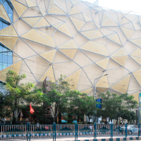 The modern building, reminiscent of architectural wonders found in India, showcases an intricate geometric facade with triangular gold and glass panels. Trees line the street in front, while traffic barriers run parallel to the sidewalk. A clear sky stretches above, dotted with some street signs nearby.