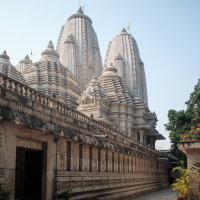 The image depicts an Indian Hindu temple complex with intricate stone carvings and towering shikharas against a clear blue sky. The exterior wall features detailed motifs, and lush greenery to the right enhances the serene atmosphere.