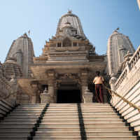 A man stands at the top of a grand staircase leading to a magnificent Hindu temple in India. The temple, made of intricately carved white stone, features multiple towering spires against a clear blue sky. The architectural details reflect traditional religious design.