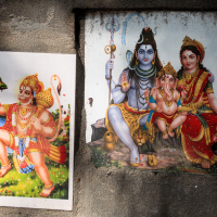 Two vibrant religious prints on a textured stone surface evoke the essence of India. Left: Hanuman holding a mountain and mace. Right: Shiva with a trident, Parvati, and Ganesha seated together, adorned in colorful traditional attire and jewelry, conveying divinity and cultural richness.