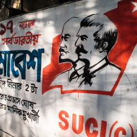 A mural on a wall showcases illustrations of two men in profile. The background features a red map shape of India and a star, with text in Bengali reading "SUCI(C)" in bold red lettering alongside event details. Shadows of trees partially overlay the mural, adding to its vibrant narrative.