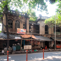 A weathered two-story building with arched windows and a faded facade stands under lush green trees typical of India. Below are small shops with makeshift tarps. A few red poles line the road in front, and a mix of light and shadow falls across the scene, indicating a sunny day.