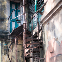 A weathered spiral staircase with ornate railings leads to a closed wooden door with peeling paint on a dilapidated building reminiscent of old India. The wall shows signs of age, with patches of faded yellow and green paint. A barred window sits beside the staircase, casting shadows in the sunlight.