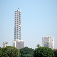 A tall skyscraper under construction with cranes on top rises majestically against a clear blue sky in India. The building is surrounded by other high-rise structures, and in the foreground, a lush green park with manicured grass hosts a few people sitting on benches.