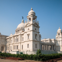 A large, ornate building made of white marble stands under India's clear blue sky. It features a central dome, multiple smaller domes, and intricate architectural details. In the foreground, there's a lawn and some construction materials. The structure exudes a sense of grandeur and history.