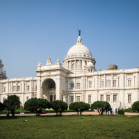 The image showcases the Victoria Memorial in Kolkata, India, a stunning white marble building with a central dome topped by a statue. Flanking it are smaller domes and towers. This iconic Indian landmark is surrounded by lush green lawns and bushes under a clear blue sky.
