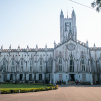 A large Gothic-style cathedral in India stands under a clear blue sky. The facade features pointed arches, ornate carvings, and a central rose window. Several people are gathered at the entrance, surrounded by lush greenery and a paved pathway in the foreground.