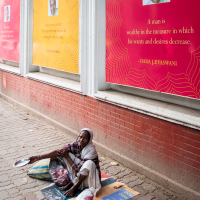 An elderly person sits on a mat on the bustling sidewalk of an Indian city, holding a bowl. Behind them, large red and yellow posters with quotes and a smiling man's photo adorn the brick wall, creating an urban backdrop. Various bags and personal belongings surround them in this vibrant scene.