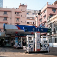 In bustling India, a city gas station with two fuel pumps stands before a pink multi-story building. Signs for "Diesel" and "Petrol" hover above the pumps, and there's a small convenience store to the left. Urban buildings and patches of greenery encircle this vibrant spot.