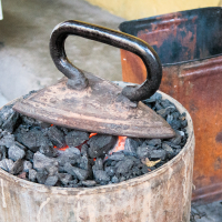 An old-fashioned coal iron rests on hot coals in a metal container, reminiscent of traditional scenes from India. Nearby, a rusted metal bin adds to the rustic setting, with concrete flooring and a yellow wall in the background. The coals glow red beneath the iron, indicating the heat used for pressing clothes.
