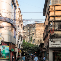 A bustling Indian urban street scene with narrow lanes, flanked by tall, weathered buildings. Shops line both sides, including a unisex beauty salon. People walk and bike through the street, and a black car is parked nearby. Overhead, many tangled electric wires cross the sky.