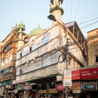 A bustling street scene in India features a multi-story building with a mosque on the upper levels, marked by its green domes and minarets. Below, the ground floor hosts various shops, including a juice center. Several people walk along the street, and some gather near scooters parked by the curb.