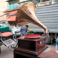 A vintage gramophone with a brass horn and wooden base sits on a table beside a plastic water bottle. In the background, an Indian rickshaw and a large truck are parked on a bustling street lined with buildings, signs, and numerous utility wires overhead.