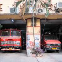 Two red fire trucks are parked inside an old fire station in India. The building has a faded exterior with peeling paint and visible air conditioning units. A small chain blocks the entrance, and a few plants are placed near the trucks, capturing a vintage and rustic ambiance.