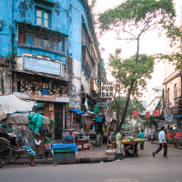 A bustling urban street scene in India depicts a vibrant corner with a blue, weathered building. People are walking; a vendor sells produce; signs indicate a pub. A parked rickshaw is nearby, and trees add greenery. The street is lined with various goods and small shops under a clear sky.