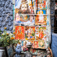 A colorful collage of Hindu deities is displayed against a textured blue wall. At the forefront, there are various religious idols and offerings from India, including a black shivling adorned with a white trident symbol. Marigold flowers and small statues surround the centerpiece.
