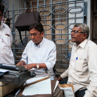 Three men are gathered around an old typewriter on a street in India. The man in the middle is typing, while the others look on. Papers and a ruler are placed on the table. They stand before a metal gate and building entrance, all dressed in light-colored, casual shirts.