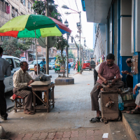 A bustling street scene in India depicts several men on a sidewalk. On the left, a man sits at a typewriter under a colorful umbrella. To the right, two men sit on steps, one with a phone. A dog lies nearby. Vehicles and buildings line the busy street, hinting at a lively urban setting.