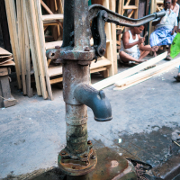 A rusty, old-fashioned hand water pump stands on a stone base in an Indian street. Behind it, several wooden frames are stacked vertically. In the background, a group of men sit on the ground, relaxing and chatting, with one wearing a traditional white dhoti. The setting is urban and informal.