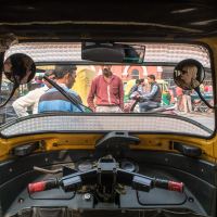 View from inside an auto rickshaw in India showing the driver's seat, handlebars, and mirrors. Outside the front, several people are standing and conversing on the street. There are additional auto rickshaws and a two-story brick building with arched windows in the background.