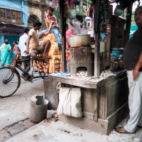 A bustling street scene in a vibrant Indian market area. A roadside vendor stands by a traditional clay stove with pots steaming. A bicyclist rides by, and a man sits on a rickshaw. The backdrop features colorful buildings and pedestrians walking past in various attire.