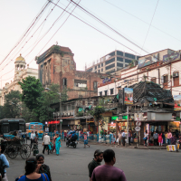 A busy street scene in Kolkata, India, with numerous pedestrians, rickshaws, and vehicles. The vibrant backdrop showcases a mix of historic and modern Indian buildings, including a large brick structure. Overhead wires crisscross the scene amidst various shop signs and billboards.